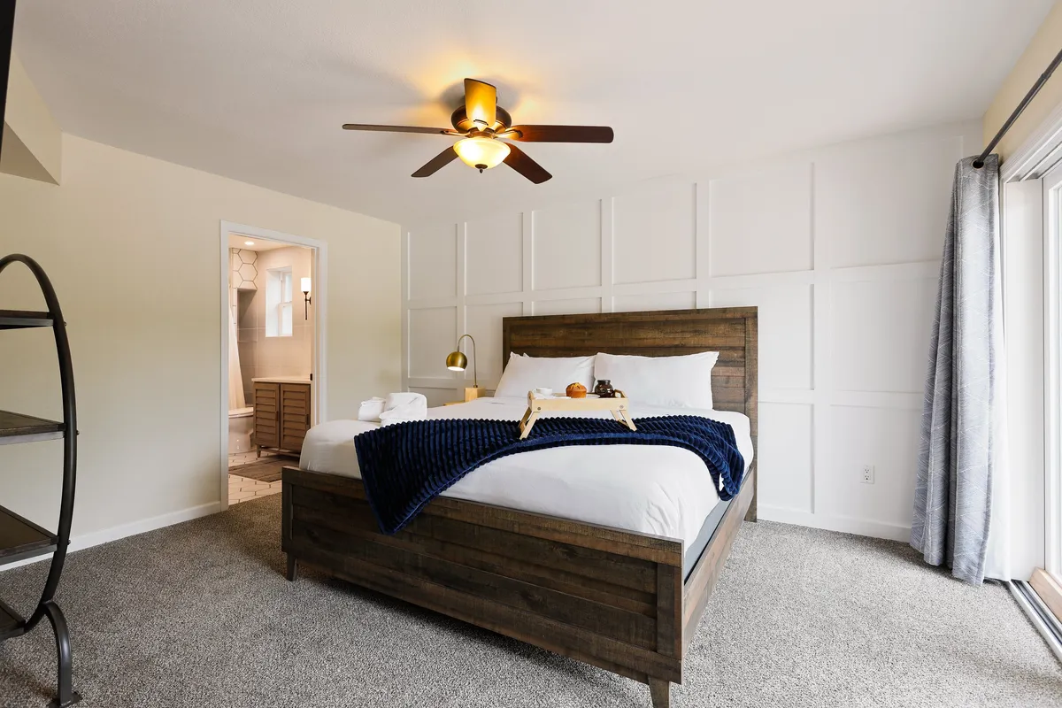 A cozy bedroom with a navy blanket and pillows, wooden nightstand, ceiling fan, and an open door to the bathroom. The ambiance is serene, with soft natural light making the space feel restful and inviting.