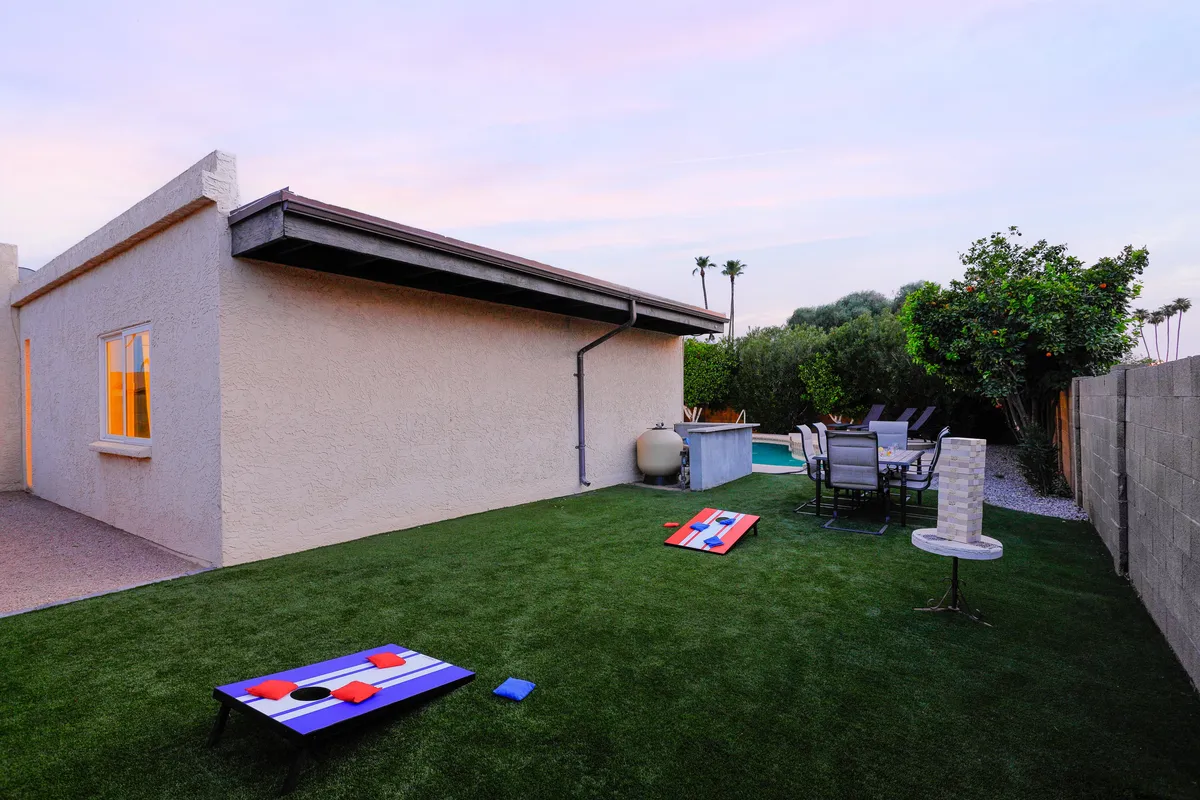 This image captures the outdoor seating area bordered by lush greenery, with lawn games like cornhole in the foreground. The patio is set up for relaxing and socializing, while the surrounding grass and plants add to the tranquil, playful mood.