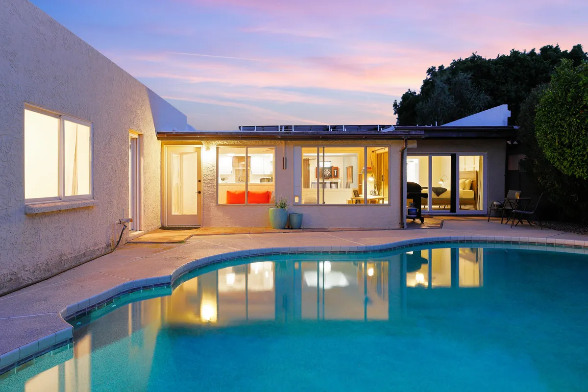A serene exterior shot featuring a sparkling swimming pool, large house windows, outdoor seating, and a dramatic sunset sky. The inviting setup hints at relaxation and downtime after work, with cozy spaces for enjoying peaceful evenings.