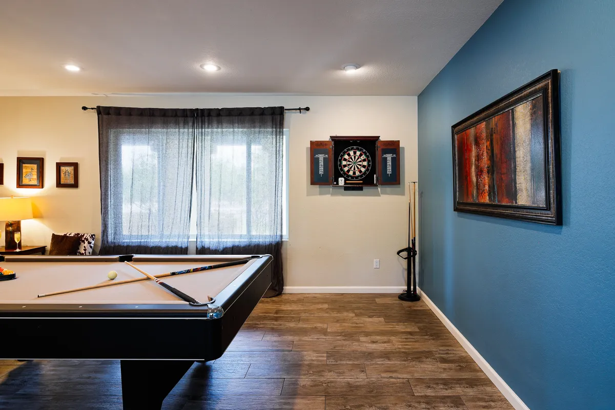 A recreational interior shot featuring a pool table, dartboard, large window flooding the space with light, framed artwork, and a cozy sofa. The scene radiates an energetic yet welcoming mood, ideal for group games and relaxing between rounds.