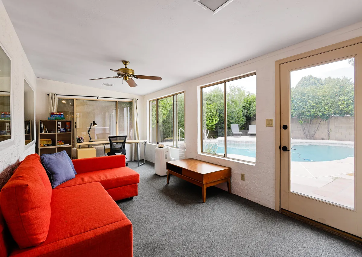 The living room scene highlights a red couch (which doubles as a sleeper sofa), a wooden coffee table, a desk with chair, and a scenic pool view through the window. The space feels inviting and multifunctional.