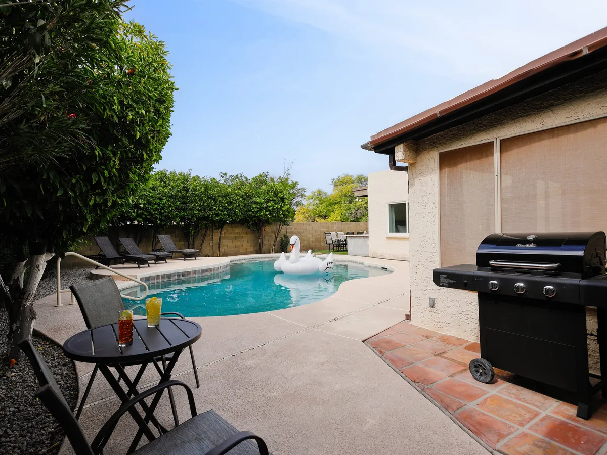 A cheerful scene showing the swimming pool with a playful swan float, a barbecue grill, and an inviting outdoor seating area. The surrounding backyard is well-kept and radiates a sense of celebration and togetherness, ideal for sunbathing, barbecuing, and poolside relaxation.