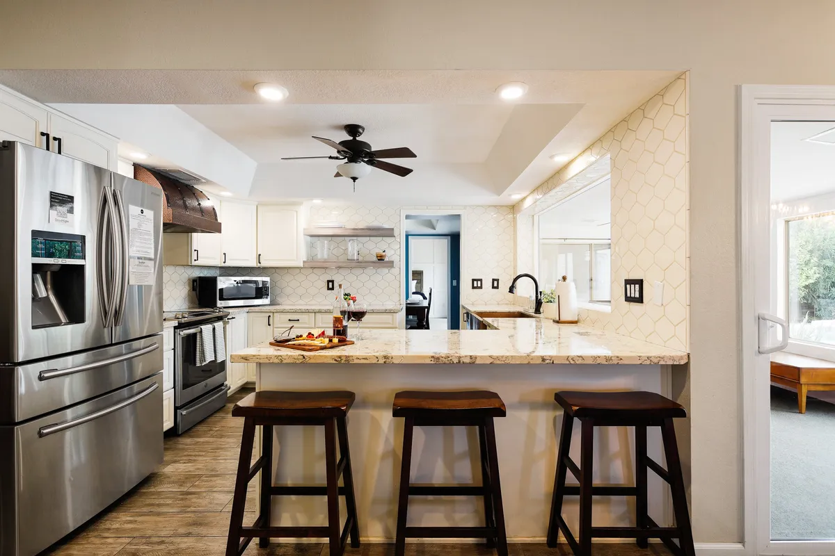 A modern kitchen with stainless steel appliances, granite countertop, bar stools at a kitchen island, and an open doorway leading to an adjacent room. The space feels welcoming and perfect for group cooking or casual meals.