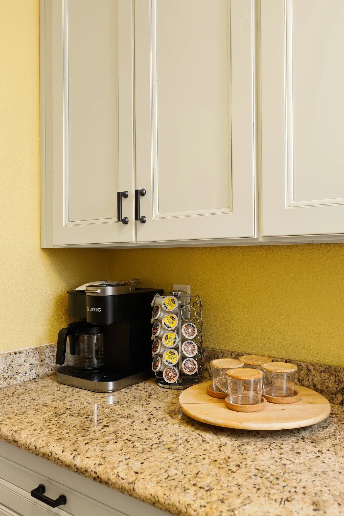 A warm, cozy corner featuring a Keurig coffee maker, coffee pod holder, and a wooden serving platter with glasses—suggesting a space for morning routines or casual conversation.