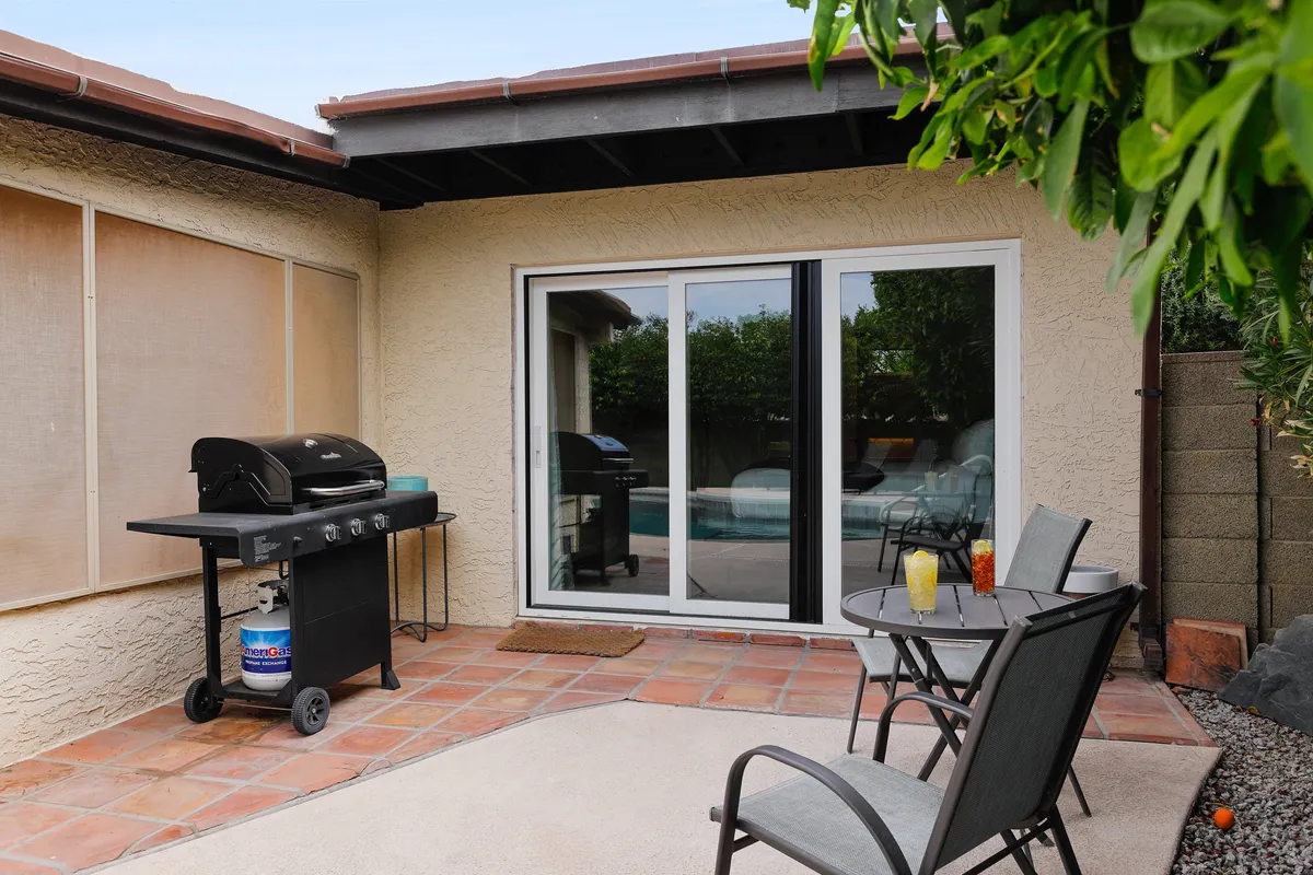 This image presents a lively backyard setting featuring a grill, patio furniture, a sliding glass door leading inside, and a swimming pool. The scene is styled for social gatherings with comfortable seating and ample space for grilling and outdoor meals. The pool and nearby patio suggest a relaxing atmosphere for friends to unwind together.