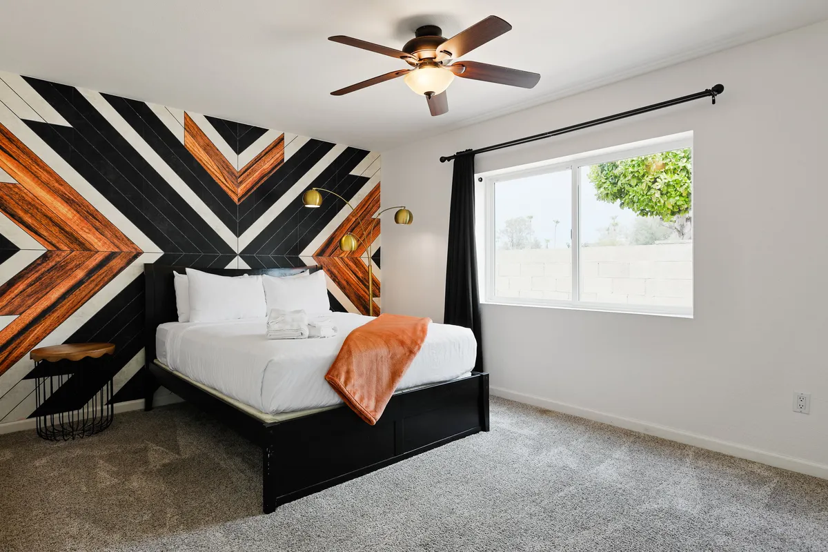 A stylish bedroom with a geometric accent wall, bed dressed in white linens and an orange throw, wooden side table, and a window letting in abundant natural light. The space feels both contemporary and restful.
