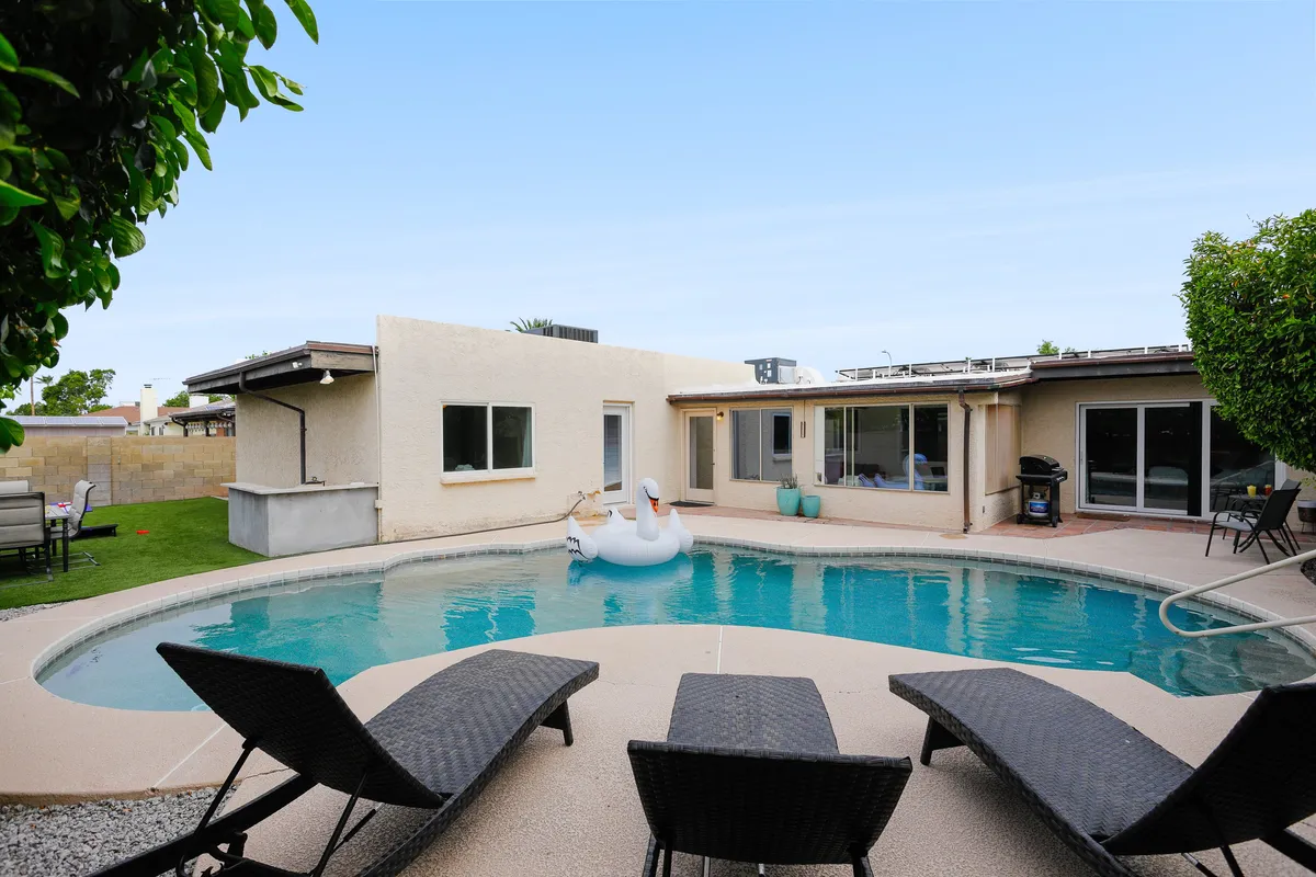 A sunny backyard scene with a sparkling swimming pool, swan float, lounge chairs, and lush greenery. The space is inviting for swimming, sunbathing, social gatherings, and outdoor dining.