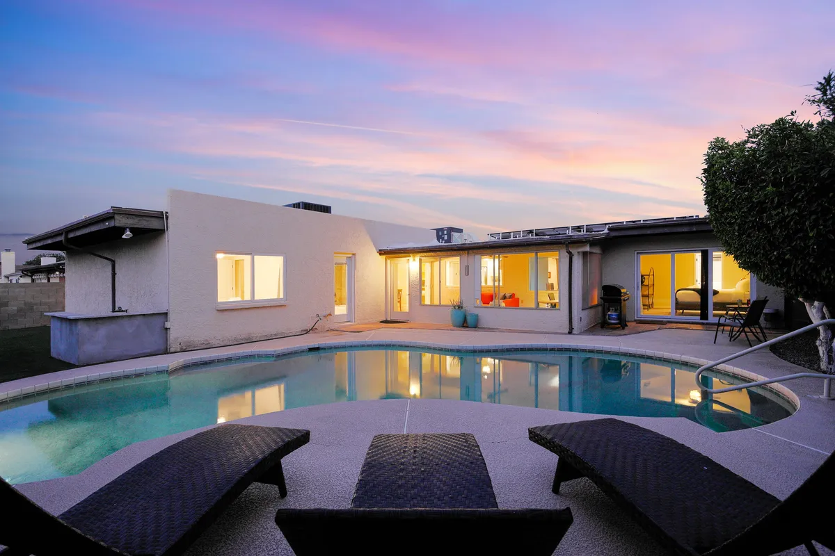The hero image showcases the inviting backyard of Pebble Retreat, featuring a sparkling swimming pool surrounded by comfortable lounge chairs, all set against the warm glow of an Arizona sunset. Large windows hint at the spacious interior, while the serene atmosphere invites guests to relax, swim, sunbathe, or enjoy outdoor dining.