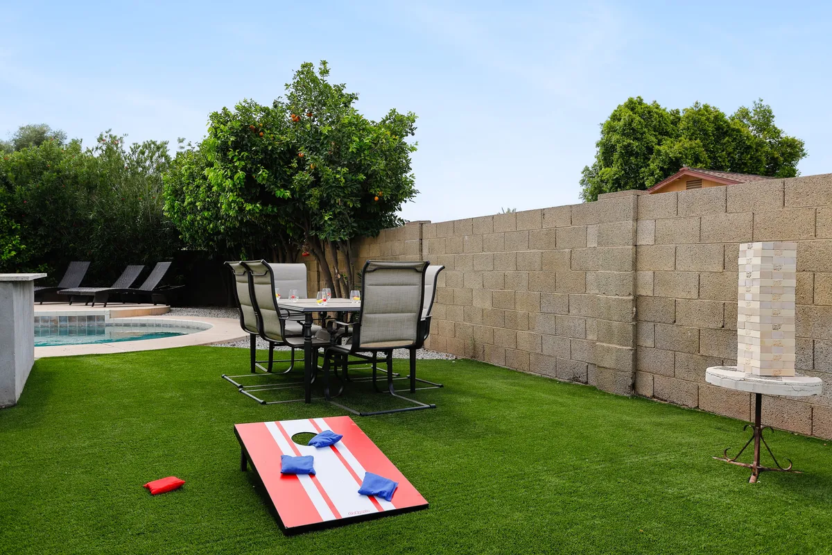 A lively backyard scene featuring the private swimming pool, green grass, lounge chairs, a cornhole lawn game set up, and an outdoor dining table with chairs. The area is bathed in natural sunlight, and the arrangement encourages both relaxation by the pool and active outdoor play.